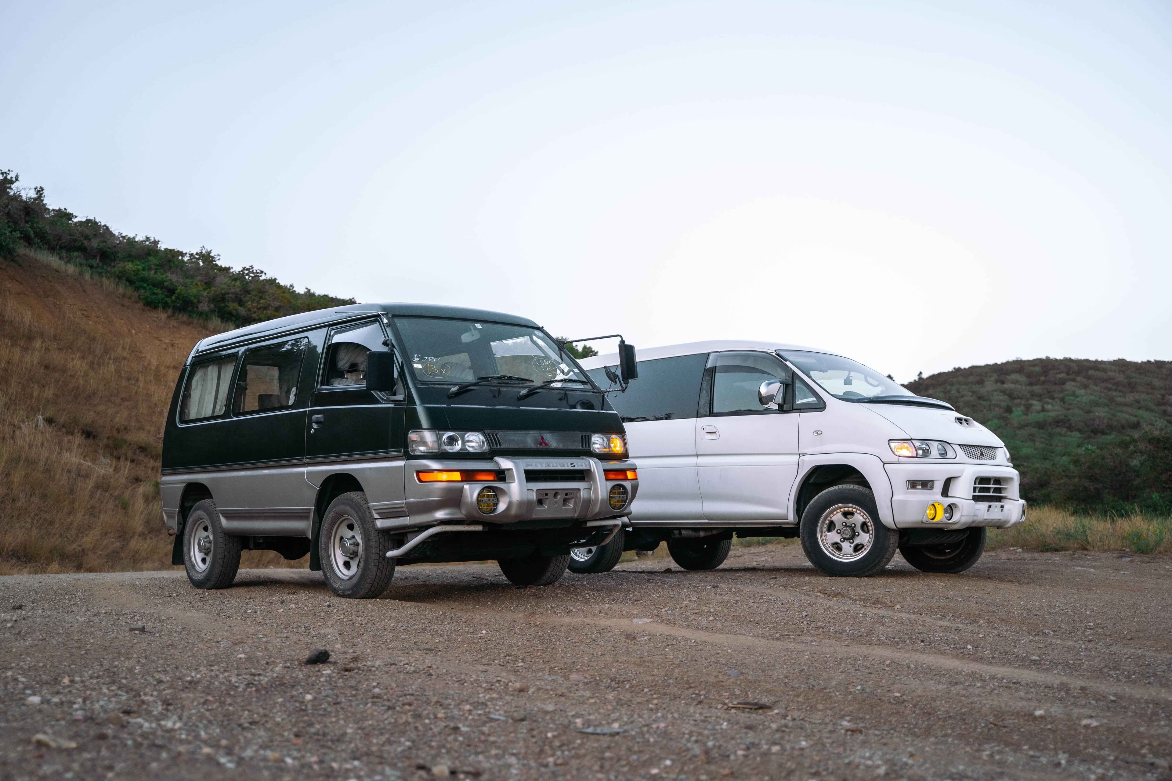 Green Mitsubishi Delica Star Wagon (left) and white Mitsubishi Delica Space Gear (right) parked side-by-side on a dirt road.