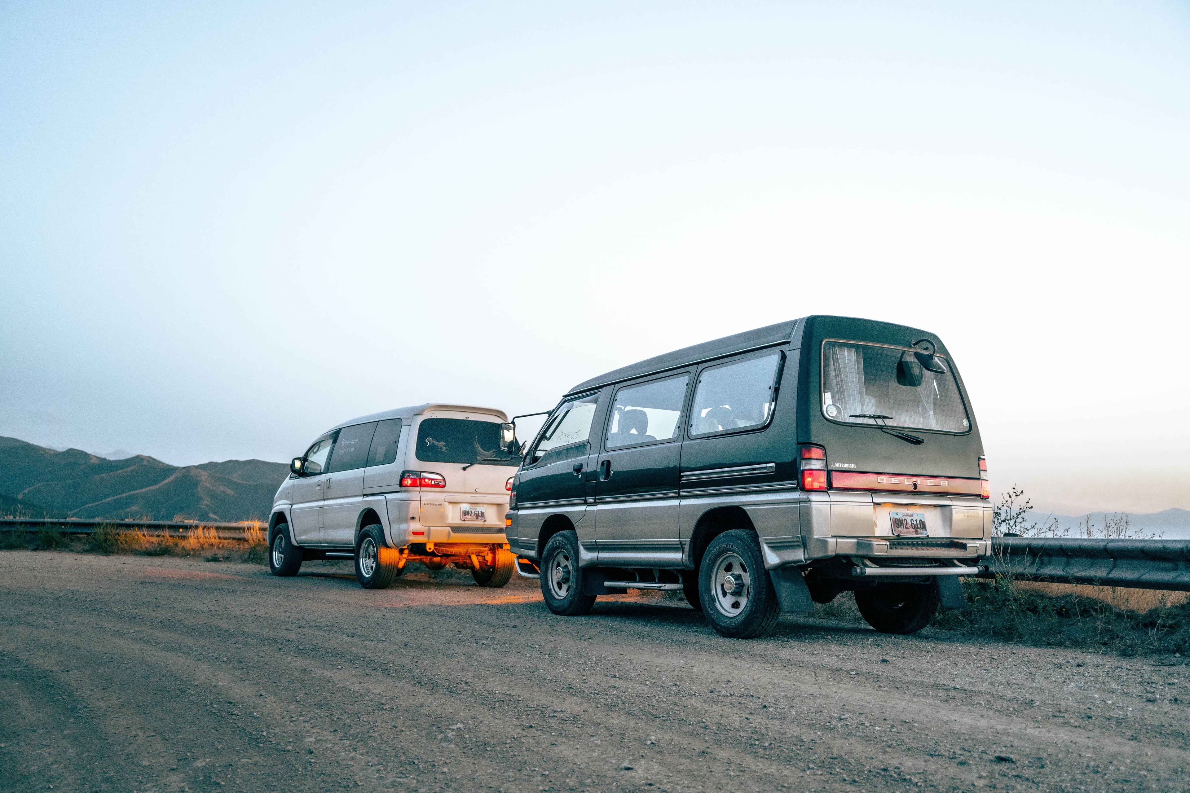 White Mitsubishi Delica Space Gear parked in front of green Mitsubishi Delica Star Wagon on a dirt road.